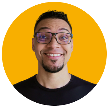Foto de Dev sorrindo, usando óculos e camisa preta, barba e fundo amarelo.